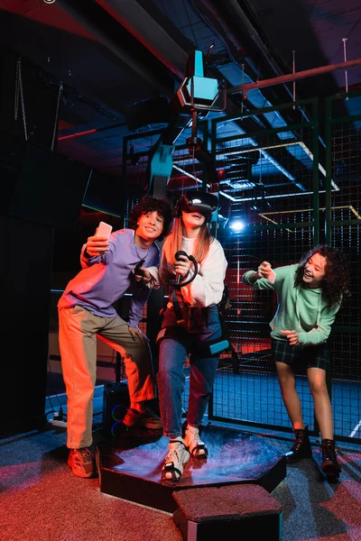 Amigos multiétnicos alegres tirando fotos perto de jogos de meninas na plataforma vr — Fotografia de Stock