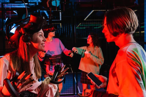 Amazed teenage girl in vr headset gesturing while talking to friend in game zone — Stock Photo