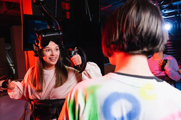 Joyeuse fille souriant près des garçons adolescents dans la zone de jeu vr, premier plan flou — Photo de stock