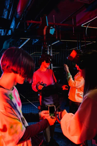 Adolescente sosteniendo teléfono inteligente con pantalla en blanco cerca de amigos en la zona de juego vr - foto de stock
