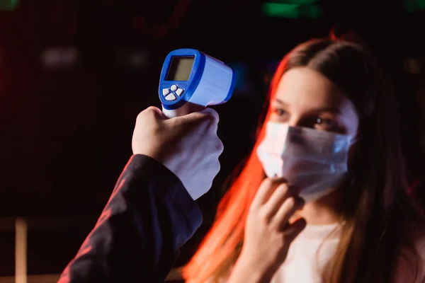 Selective focus of pyrometer in hand of controller near blurred teenage girl in safety mask — Stock Photo