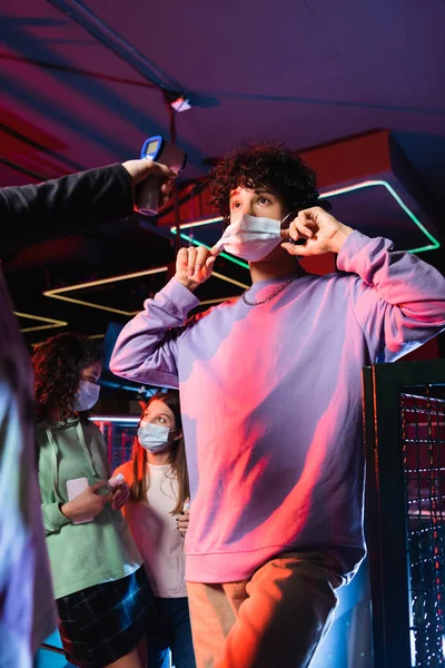 Teenager putting on protective mask near controller with pyrometer and interracial friends — Stock Photo