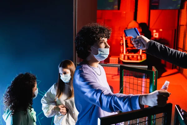 Подростковый геймер в медицинской маске держит антисептик для рук рядом с контроллером с пирометром и межрасовыми друзьями — стоковое фото