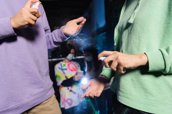 Мультиэтнические друзья, применяющие средства для дезинфекции рук вблизи размытых геймеров в медицинских масках — стоковое фото