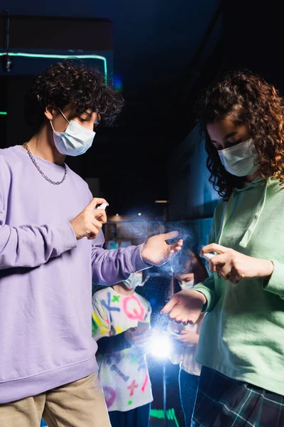 Adolescentes multiétnicos en máscaras médicas aplicando desinfectante de manos en el club de juego - foto de stock