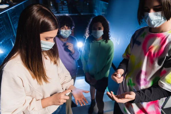 Interracial friends in medical masks spraying hand sanitizer near entrance of game club — Stock Photo