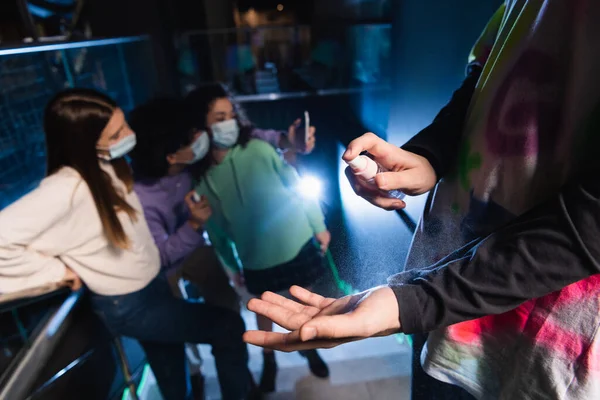 Adolescente aplicando desinfectante de manos cerca de amigos multiétnicos en máscaras de seguridad tomando selfie sobre fondo borroso - foto de stock