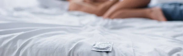 Cropped view of condom on bedding near blurred couple, banner — Stock Photo