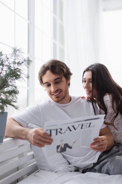 Uomo sorridente leggere giornale di viaggio vicino fidanzata sul letto — Foto stock