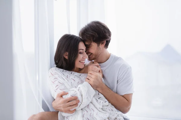 Sonriente hombre abrazando novia cerca de la cortina en casa - foto de stock