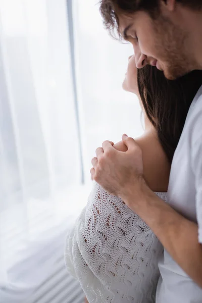 Sonriente hombre abrazando novia en suéter de punto cerca de la cortina - foto de stock