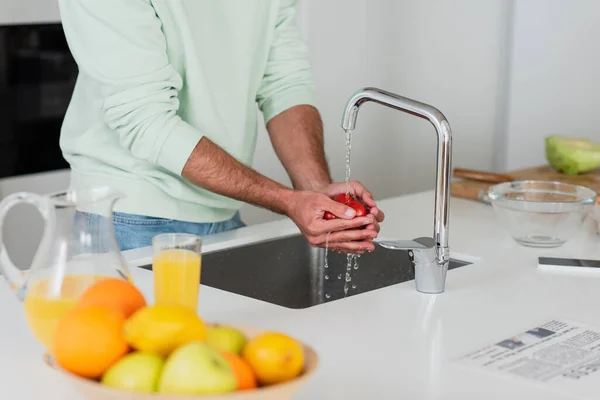 Частичный вид человека, стирающего помидоры черри рядом с фруктами и апельсиновым соком на кухне — стоковое фото