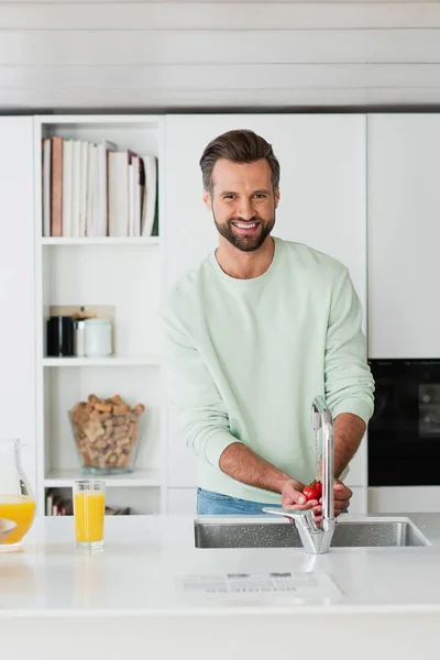 Hombre sonriendo a la cámara mientras lava tomates cherry en la cocina - foto de stock