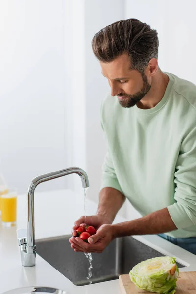 Hombre lavando tomates cherry maduros cerca de lechuga fresca en la cocina - foto de stock