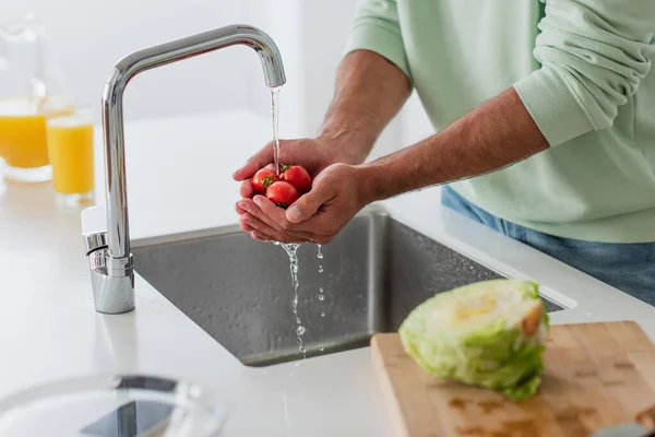Обрізаний вид людини, що миє стиглі вишневі помідори біля свіжого салату на кухні — стокове фото