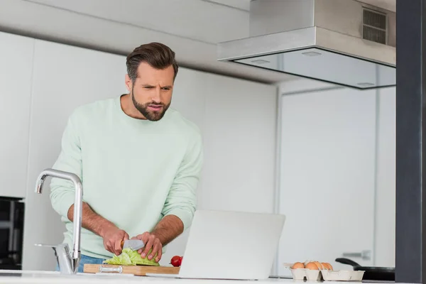 Hombre cortar verduras mientras se prepara el desayuno cerca de la computadora portátil en la cocina - foto de stock