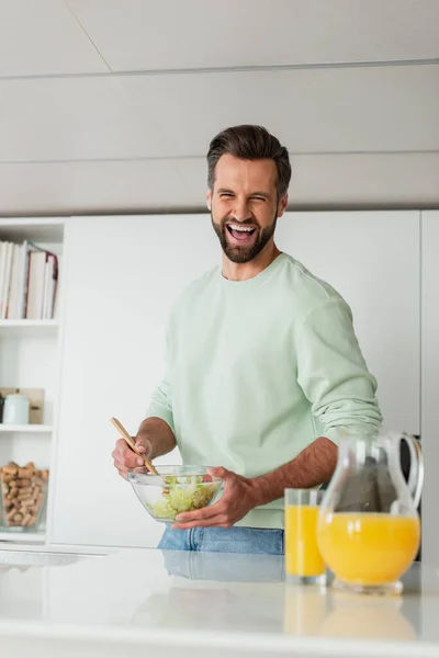 Uomo ridente mescolando insalata di verdure fresche vicino a succo d'arancia in cucina — Foto stock