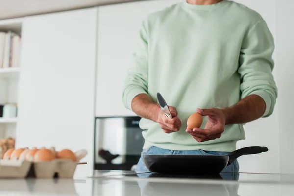 Cropped view of man holding knife and chicken egg near frying pen, blurred foreground — Stock Photo