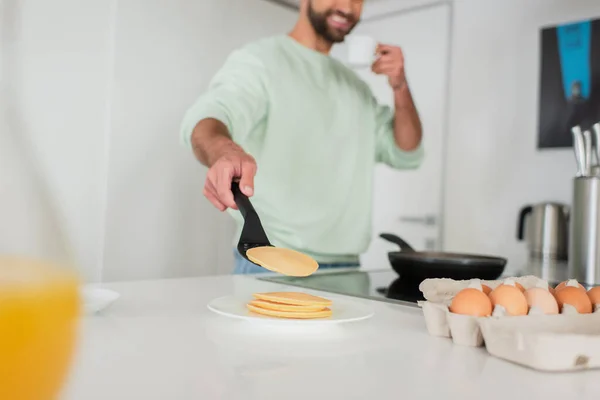 Vista recortada de hombre borroso sonriente sosteniendo taza de café mientras cocina panqueques - foto de stock