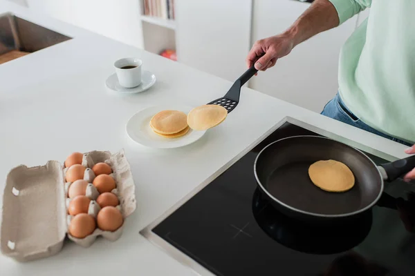 Partial view of man cooking pancakes near chicken eggs and cup of tea in kitchen — Stock Photo