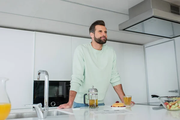 Hombre positivo mirando lejos cerca de té verde y deliciosos panqueques en la cocina - foto de stock