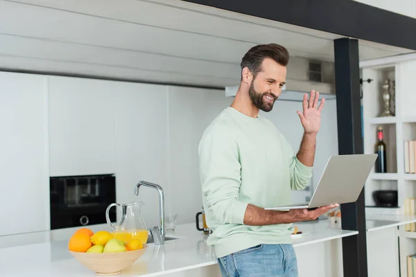 Щасливий чоловік махає рукою під час відеодзвінка на ноутбук на кухні — Stock Photo
