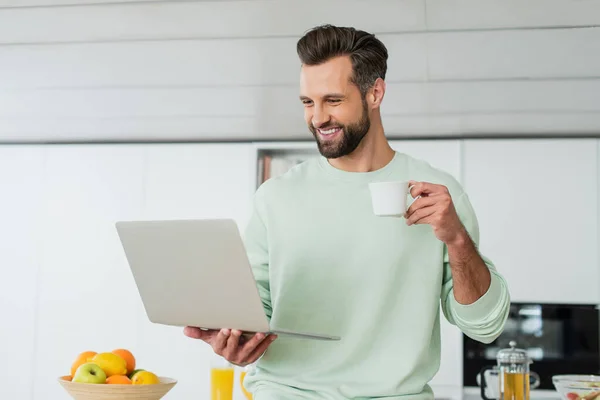 Homem feliz olhando para laptop enquanto bebe café na cozinha — Fotografia de Stock