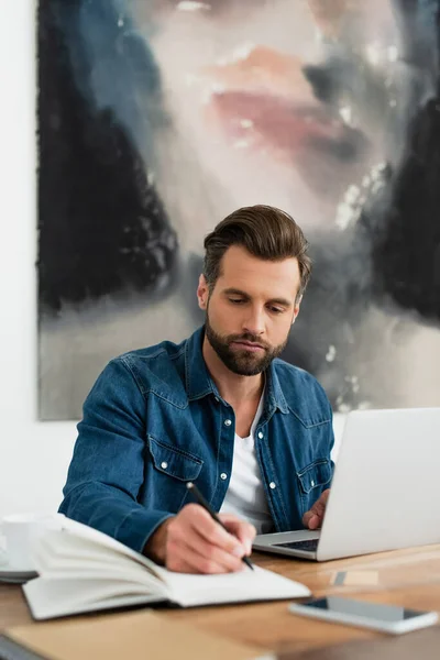 Hombre con camisa de mezclilla escribiendo en cuaderno mientras trabaja en casa - foto de stock