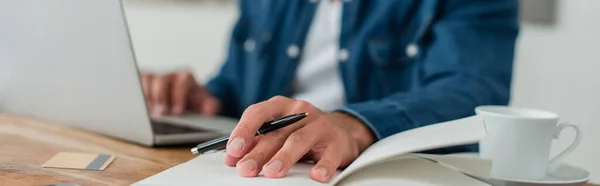 Partial view of blurred freelancer holding pen while working at laptop, banner — Stock Photo