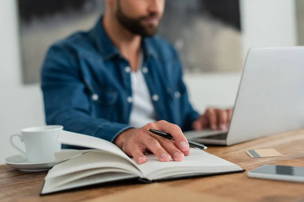 Ausgeschnittene Ansicht eines Telearbeiters, der in der Nähe von Notebook und Laptop auf verschwommenem Hintergrund arbeitet — Stockfoto