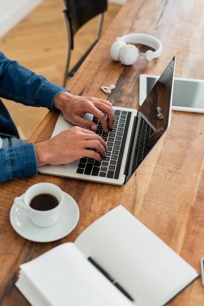 Vista recortada del freelancer escribiendo en el ordenador portátil cerca de la tableta digital y la taza de café - foto de stock