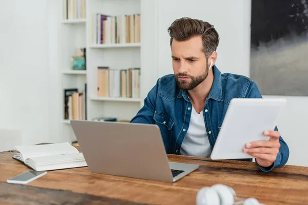 Serious man looking at laptop while holding digital tablet — Stock Photo