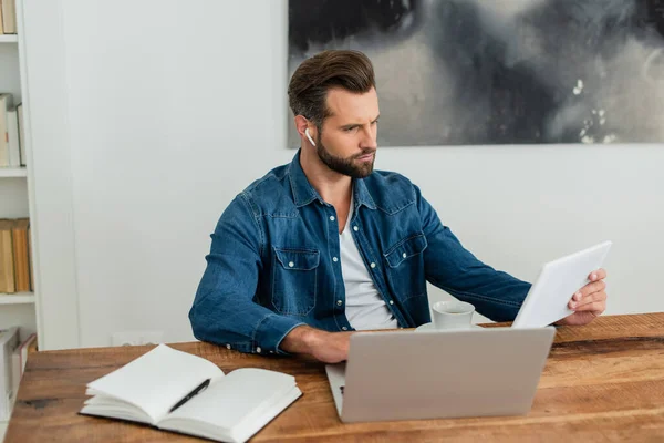 Pensativo freelancer celebración de tableta digital mientras trabaja cerca de la computadora portátil - foto de stock