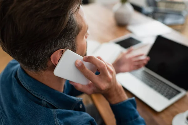 Telearbeiter spricht auf Smartphone und zeigt mit der Hand auf verschwommenen Laptop — Stockfoto