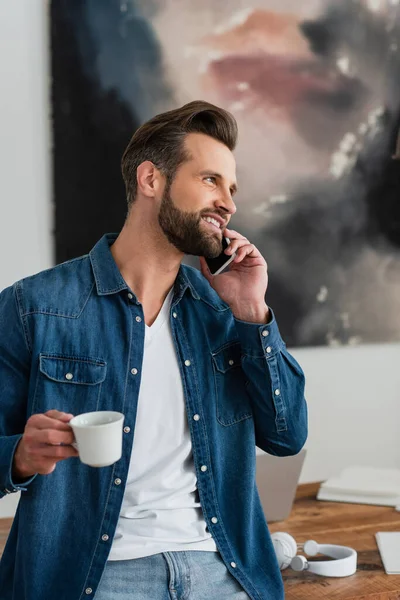 Télétravailleur heureux tenant tasse de café tout en parlant sur téléphone mobile — Photo de stock