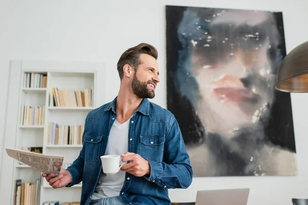 Sonriente hombre con taza de café y periódico mirando a casa - foto de stock