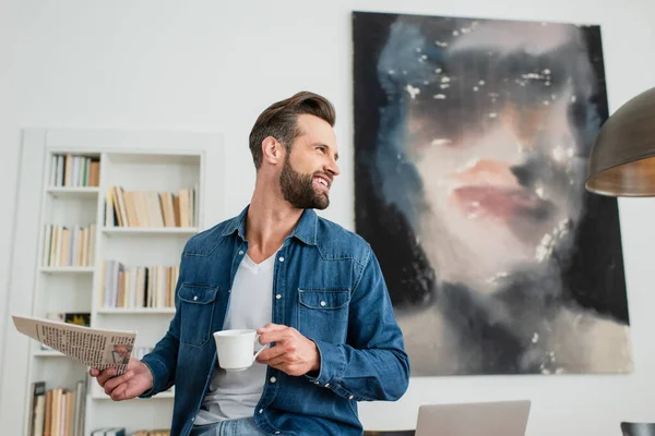 Uomo sorridente con tazza di caffè e giornale guardando lontano a casa — Foto stock