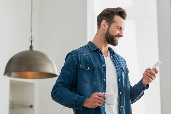 Hombre alegre con taza de café sonriendo mientras chatea en el teléfono inteligente - foto de stock