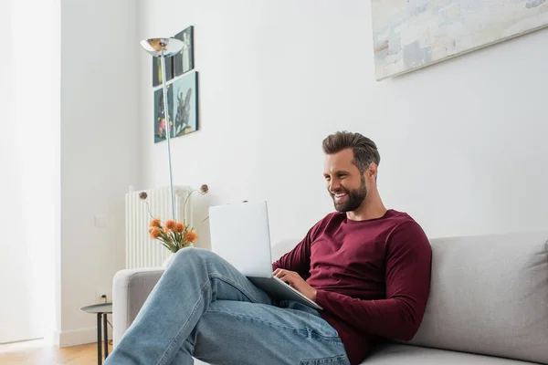 Lächelnder Mann sitzt auf Sofa und tippt auf Laptop — Stockfoto