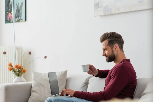 Homme tenant une tasse de café tout en étant assis sur le canapé avec ordinateur portable — Photo de stock