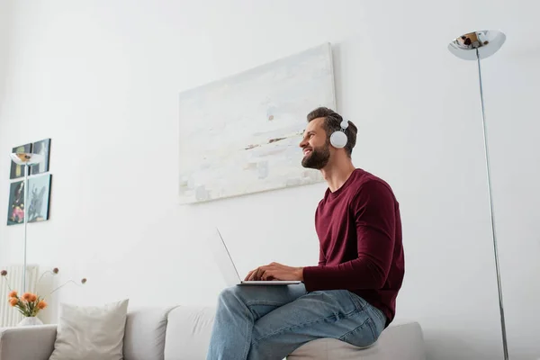 Hombre feliz en auriculares mirando hacia otro lado mientras está sentado en el sofá con el ordenador portátil - foto de stock