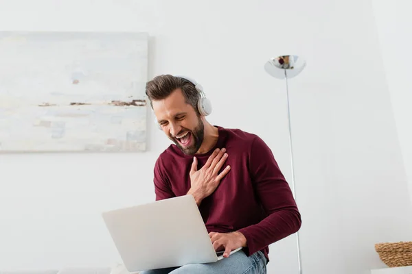 Hombre alegre en auriculares riendo cerca de la computadora portátil en casa - foto de stock