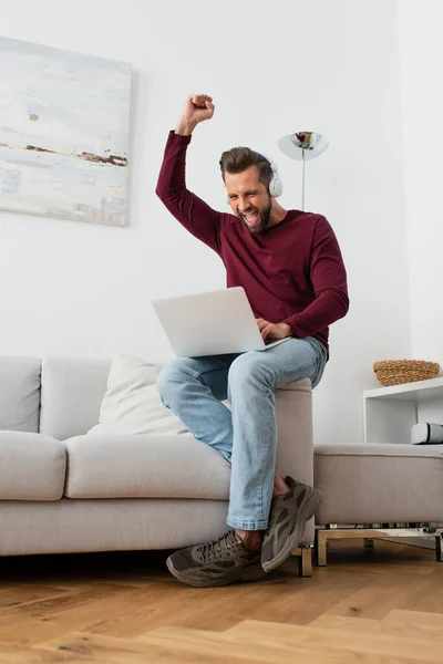 Excited man in headphones showing win gesture while looking at laptop — Stock Photo