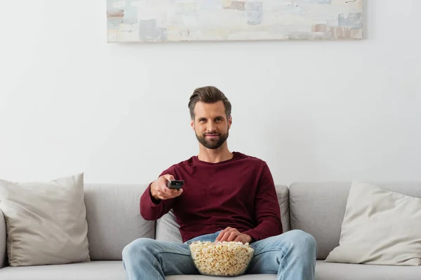 Sonriente hombre con tazón de palomitas de maíz viendo la televisión en el sofá en casa - foto de stock