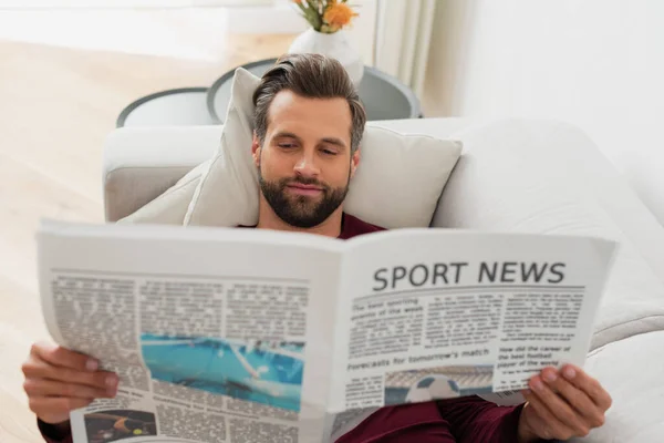 Piacere all'uomo leggere notizie sportive sul divano di casa — Foto stock