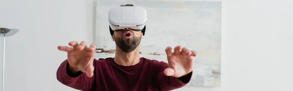 Uomo stupito gesticolando in auricolare vr mentre giocava a casa, banner — Foto stock