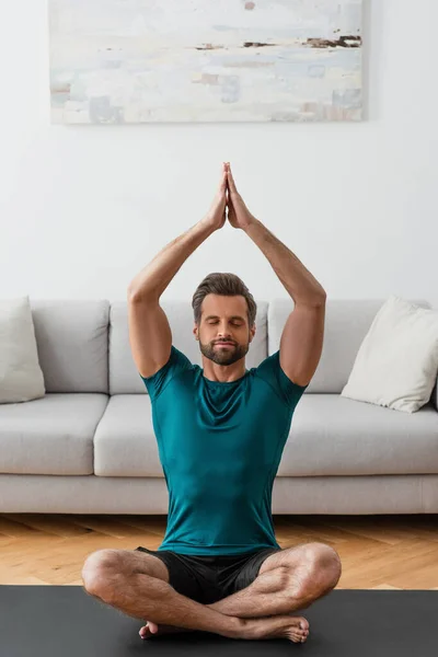 Hombre con las manos orantes y los ojos cerrados meditando en la pose de loto en casa - foto de stock