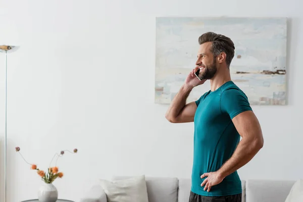 Seitenansicht eines fröhlichen Mannes, der mit der Hand auf der Hüfte steht und mit dem Handy spricht — Stockfoto