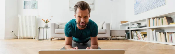 Frunciendo el ceño entrenamiento del hombre en la pose de tablón cerca de la computadora portátil, bandera - foto de stock