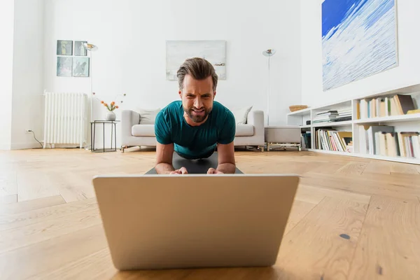 Uomo accigliato che esercita in posa tavola sul pavimento vicino computer portatile — Foto stock